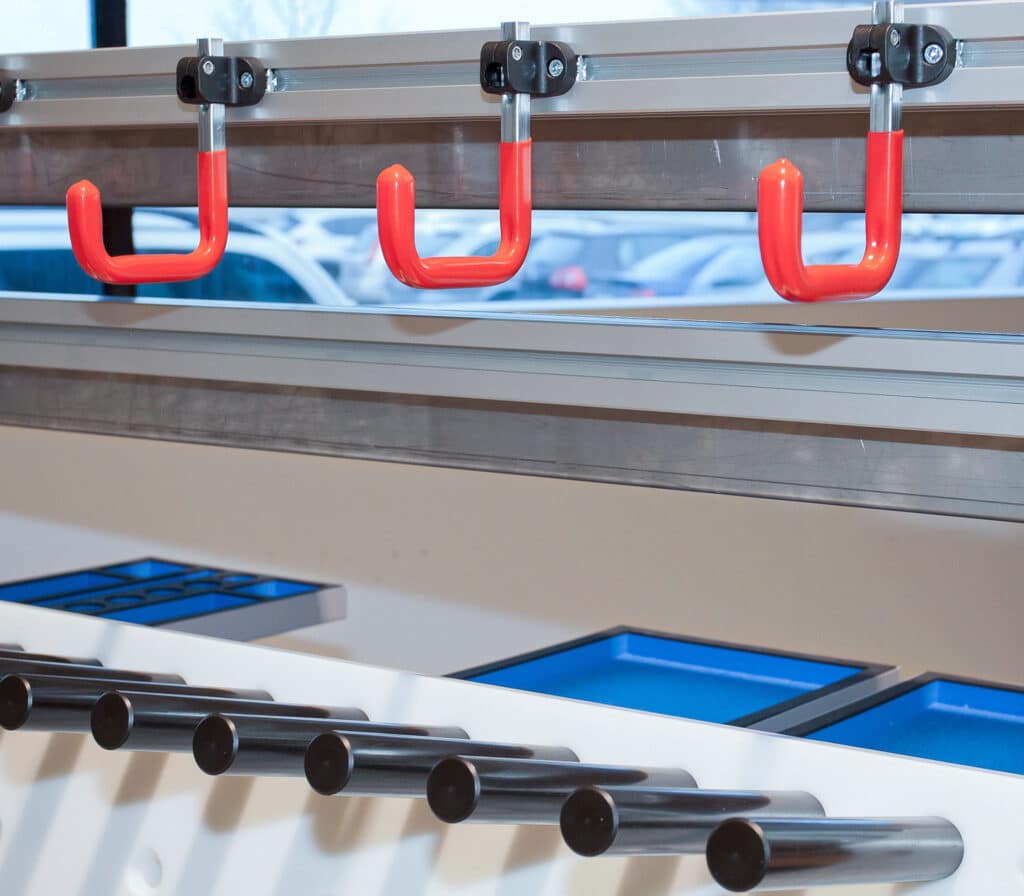 Detailansicht eines modularen Erweiterungselements von Robotunits mit roten Haken, die in einer 14 mm Nut befestigt sind. Die Anbaukomponenten lassen sich flexibel und werkzeuglos an das Profilsystem anbringen und ermöglichen eine übersichtliche Hängeaufbewahrung für Werkzeuge oder Bauteile – ideal für individuell anpassbare Arbeitsplätze.